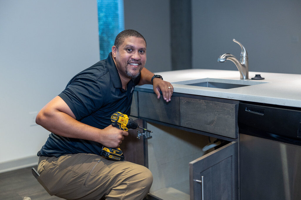 A smiling service technician kneeling at an open sink cabinet with a drill, on a residential job